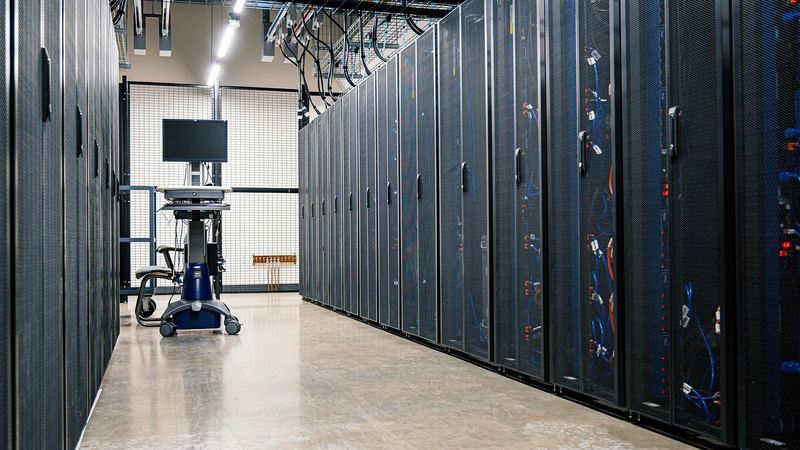 Gasse zwischen verschlossenen Serverschränken mit Gittertüren, am Ende ein Rollwagen mit Monitor